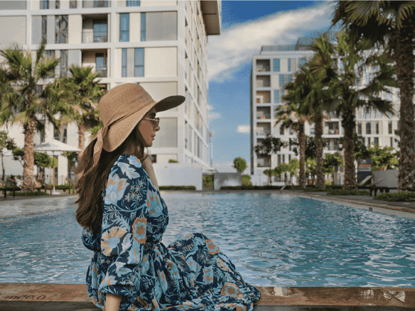 A woman is sitting on the edge of a swimming pool at Jannat by Deyaar at Dubai Production City. The pool is surrounded by lush greenery and palm trees, and the background shows a modern building.