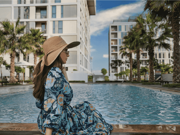 A woman is sitting on the edge of a swimming pool at Jannat by Deyaar at Dubai Production City. The pool is surrounded by lush greenery and palm trees, and the background shows a modern building.